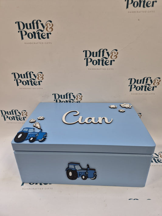 A light blue wooden keepsake box with a lid, personalised with the name' in raised silver letters on top. The box is decorated with blue tractor illustrations and small silver cloud embellishments.