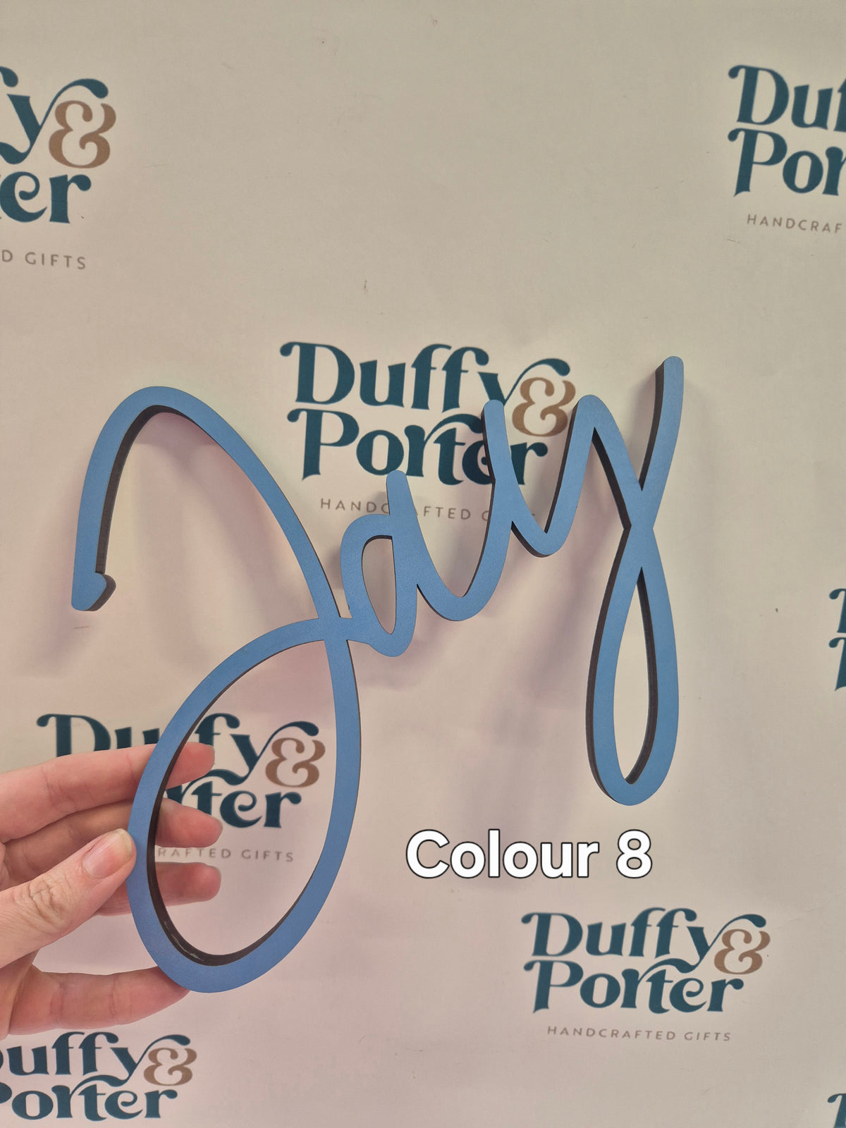 A hand holds a light blue 3D decorative script wall sign spelling 'Jay' against a white background with repeated 'Duffy & Porter' logos and a text overlay reading 'Colour 8'.
