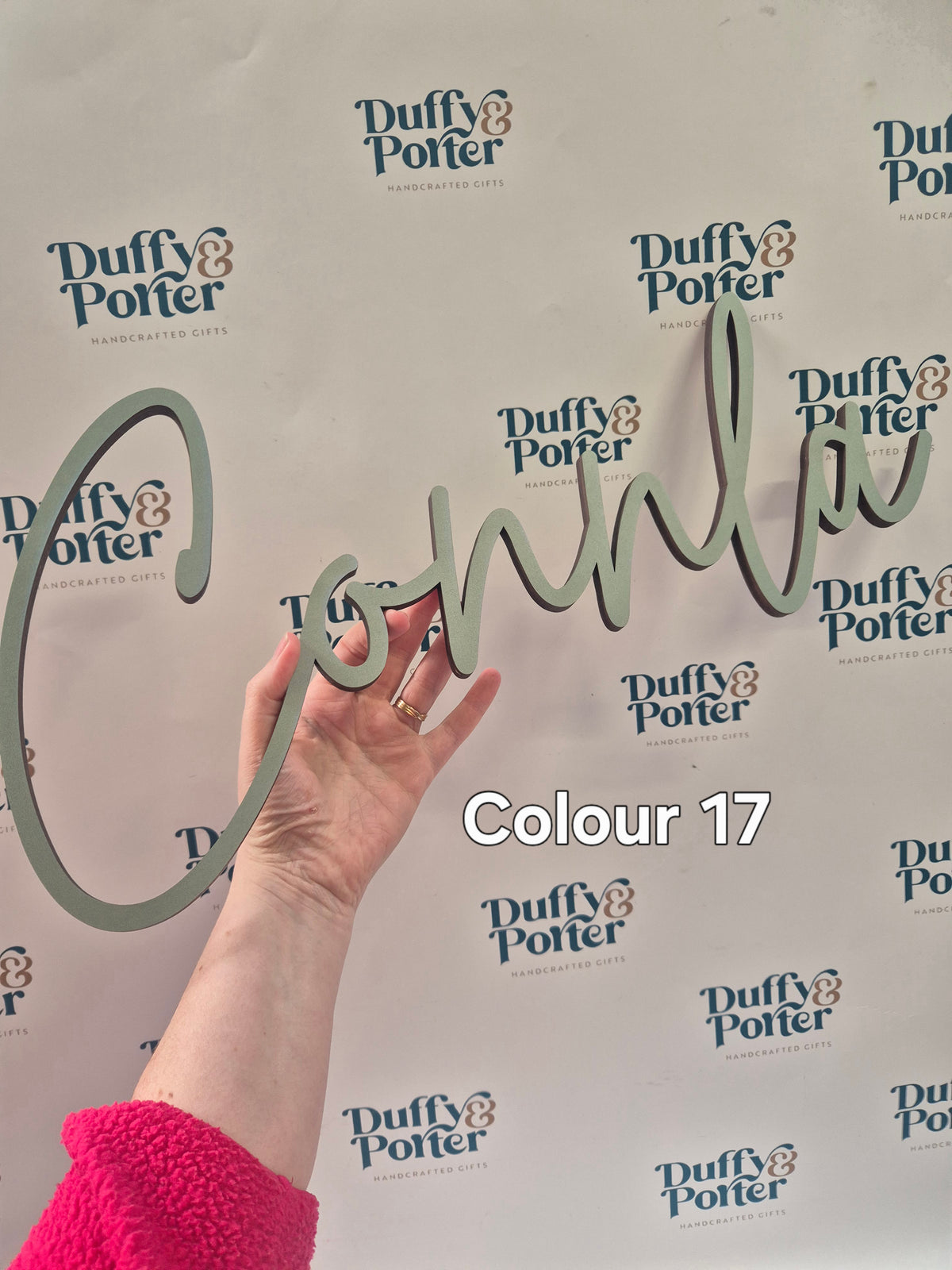 A hand holds a large, cursive, three-dimensional personalised wall name in a muted sage green color against a white background printed with the 'Duffy & Potter' logo. The text 'Colour 17' is overlaid on the image.