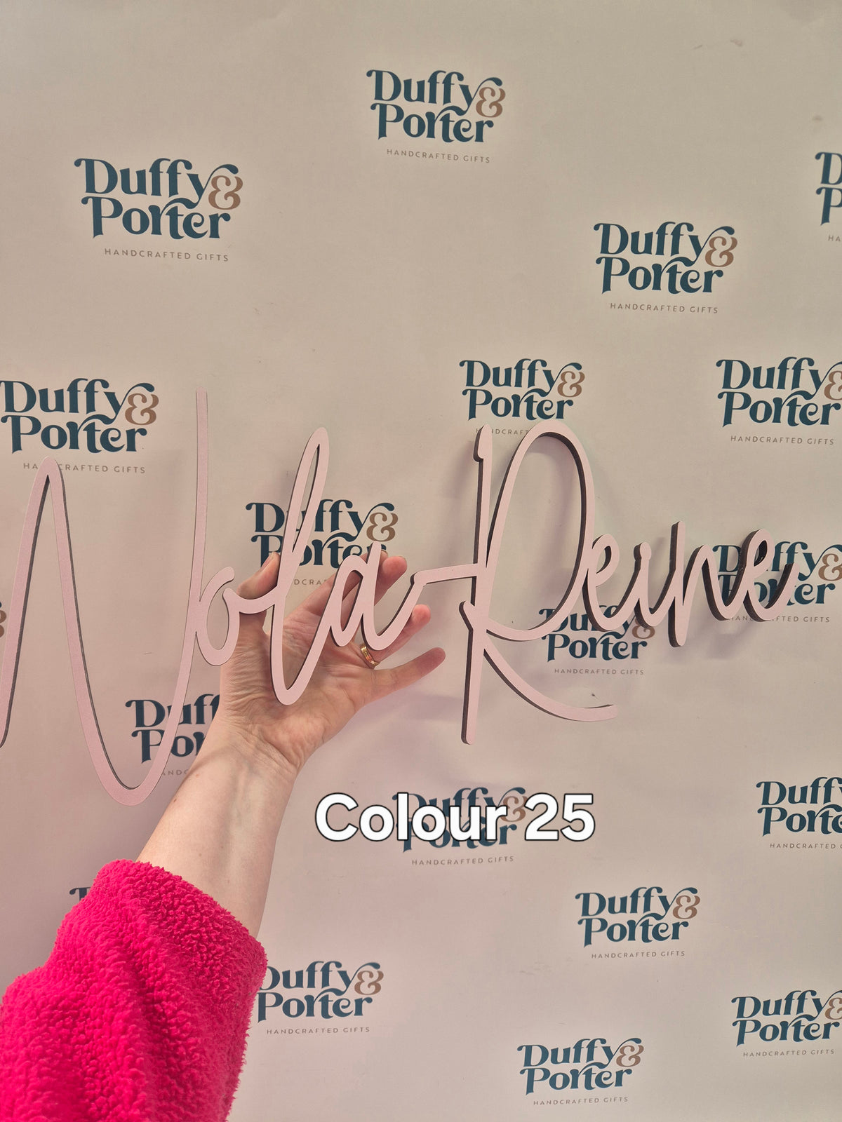 A hand holds a light pink cursive wall sign personalised with a childs name against a white background with repeated 'Duffy & Porter' logos. a text overlay reads 'Colour 25'.