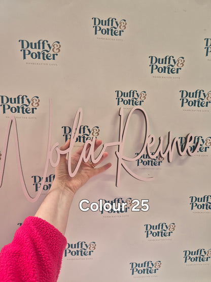 A hand holds a light pink cursive wall sign personalised with a childs name against a white background with repeated 'Duffy & Porter' logos. a text overlay reads 'Colour 25'.