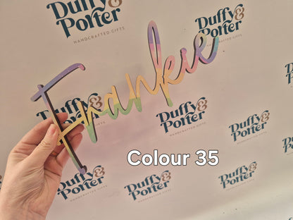 A hand holds a script wall name in the Heather font, Irish made by Duffy and Porter with a rainbow gradient finish, set against a branded white background featuring the 'Duffy & Potter' logo repeated, with 'Colour 35' text overlay.